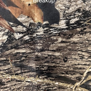 May include: Close-up of a fabric with a camouflage pattern. The design features a blend of brown, tan, and black, mimicking the appearance of tree bark and leaves. The text "Mossy Oak New Breakup" is visible at the top.