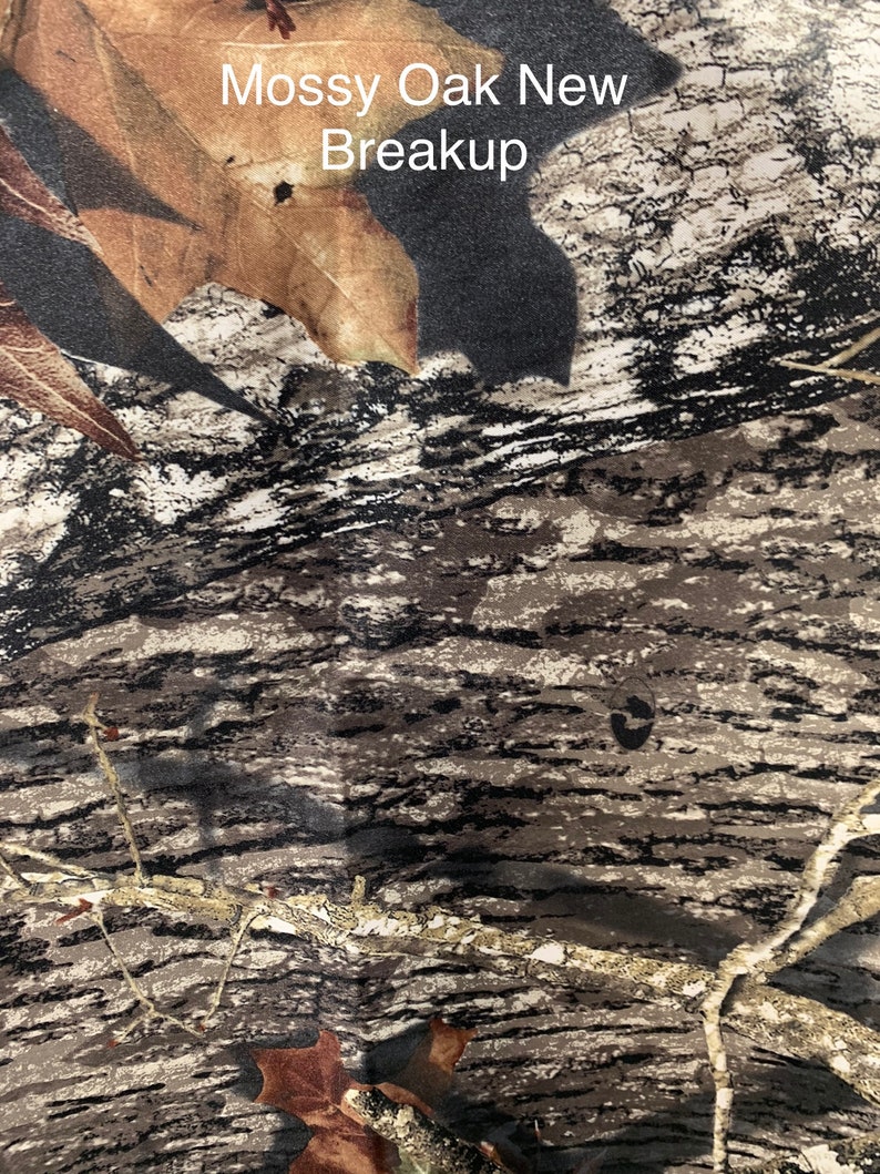 May include: Close-up of a fabric with a camouflage pattern. The design features a blend of brown, tan, and black, mimicking the appearance of tree bark and leaves. The text "Mossy Oak New Breakup" is visible at the top.
