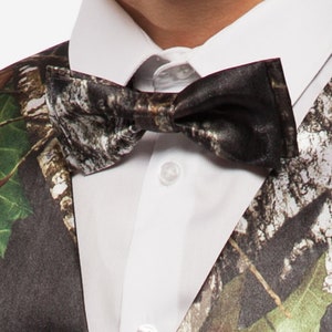 May include: A close-up of a camouflage bow tie and vest over a white dress shirt. The bow tie and vest feature a pattern of brown, green, and white, resembling a forest. The shirt has a classic collar and button closure.
