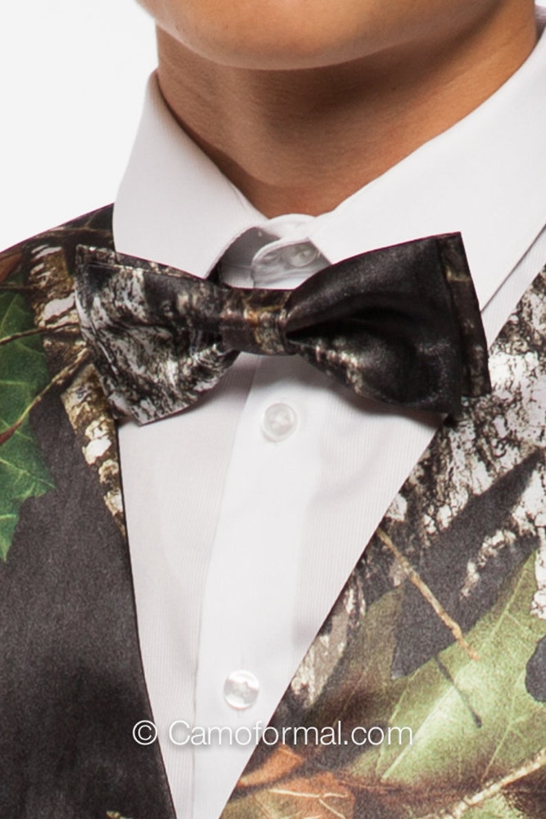 May include: A close-up of a camouflage bow tie and vest over a white dress shirt. The bow tie and vest feature a pattern of brown, green, and white, resembling a forest. The shirt has a classic collar and button closure.
