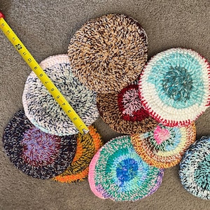 May include: A collection of nine crocheted coasters in various colors and patterns, including brown, white, blue, red, and pink. The coasters are circular and have a textured surface.