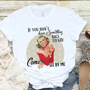 May include: White t-shirt with a vintage-style illustration of a woman in a red dress. The text on the shirt reads "If you don't have something nice to say, come sit by me."