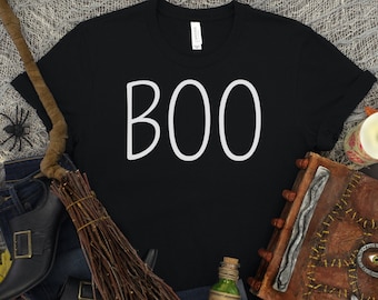 May include: Black t-shirt with white text that says "BOO" on it. The shirt is surrounded by Halloween decorations, including a broom, a book, a bottle, and some fall leaves.