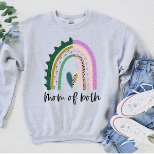 May include: A grey sweatshirt featuring a rainbow design and the text "Mom of both". The rainbow is made up of various colours and patterns, including green, yellow, pink, and leopard print.