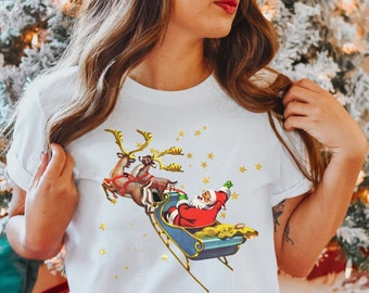 Op de afbeelding: Wit T-shirt met een vintage illustratie van Sinterklaas die in een slee vliegt die door rendieren wordt getrokken. De illustratie is omringd door gouden sterren.