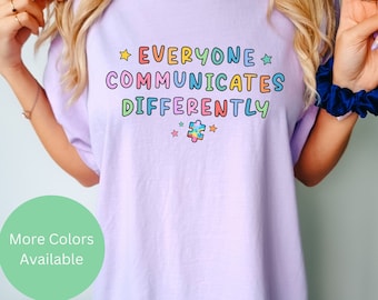 Puede incluir: Una camiseta lavanda con el texto "Everyone Communicates Differently" en colores arcoíris. Una pieza de rompecabezas colorida está debajo del texto. El texto "More Colors Available" está en un círculo verde en el lado izquierdo de la imagen. El texto "Orchid" está en cursiva negra en el lado derecho de la imagen.