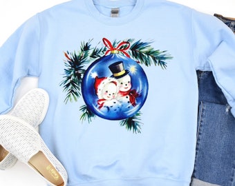 Op de afbeelding: Een lichtblauw sweatshirt met een vintage kerstontwerp met twee sneeuwpoppen in een blauwe kerstbal. De kerstbal is omringd door dennentakken.