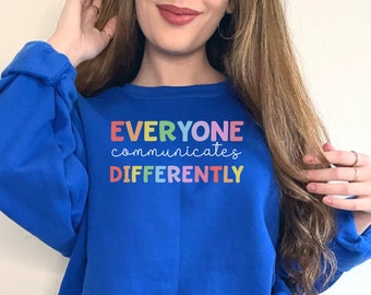 Puede incluir: Una sudadera azul real con el texto "EVERYONE communicates DIFFERENTLY" en colores arcoíris.