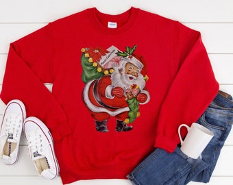 Op de afbeelding: Een rode sweatshirt met een vintage illustratie van de kerstman met cadeaus en een kous. De tekst "Merry Christmas" is zichtbaar op een label. Witte sneakers, een blauwe spijkerbroek en een witte mok staan ook op de foto.