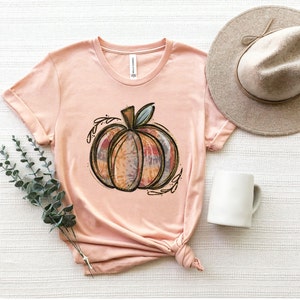 May include: A peach-colored t-shirt with a tie-dye pumpkin design. The pumpkin is outlined in gold and has a brown stem. The shirt is made of a soft, comfortable material.