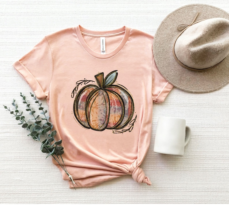 May include: A peach-colored t-shirt with a tie-dye pumpkin design. The pumpkin is outlined in gold and has a brown stem. The shirt is made of a soft, comfortable material.