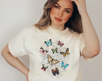 Peut inclure: T-shirt crème avec un motif de papillons colorés. Le graphisme présente diverses espèces de papillons bleus, jaunes, rouges et verts, sur fond de dessins floraux. Le t-shirt a des manches courtes et une coupe décontractée.