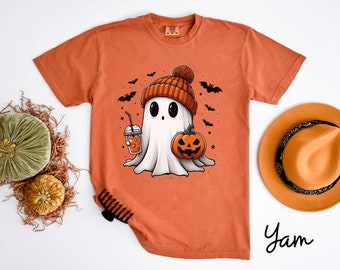May include: A burnt orange t-shirt with a cute cartoon ghost wearing a knitted hat and holding a pumpkin and a cup of coffee. The ghost is surrounded by bats and stars. The text "Yam" is printed on the bottom right corner of the shirt.