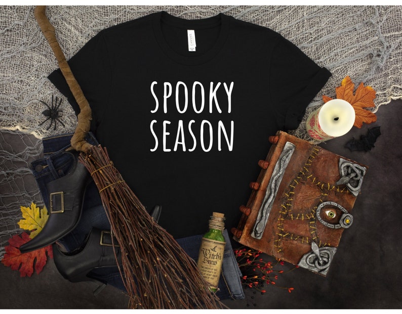 May include: Black t-shirt with white text that reads "Spooky Season". The shirt is surrounded by Halloween-themed props, including a broom, a book, and a bottle of potion.