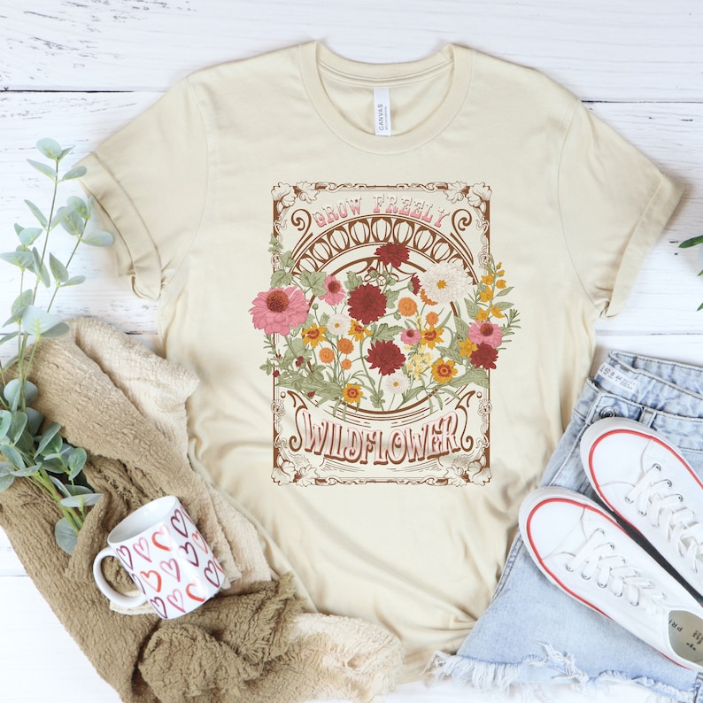 May include: A beige t-shirt with a vintage-style graphic featuring a bouquet of colorful flowers and the text "Grow Freely Wildflower".