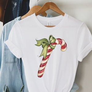 May include: White t-shirt with a red and white candy cane with a green bow on top. The candy cane is drawn in a cartoon style.