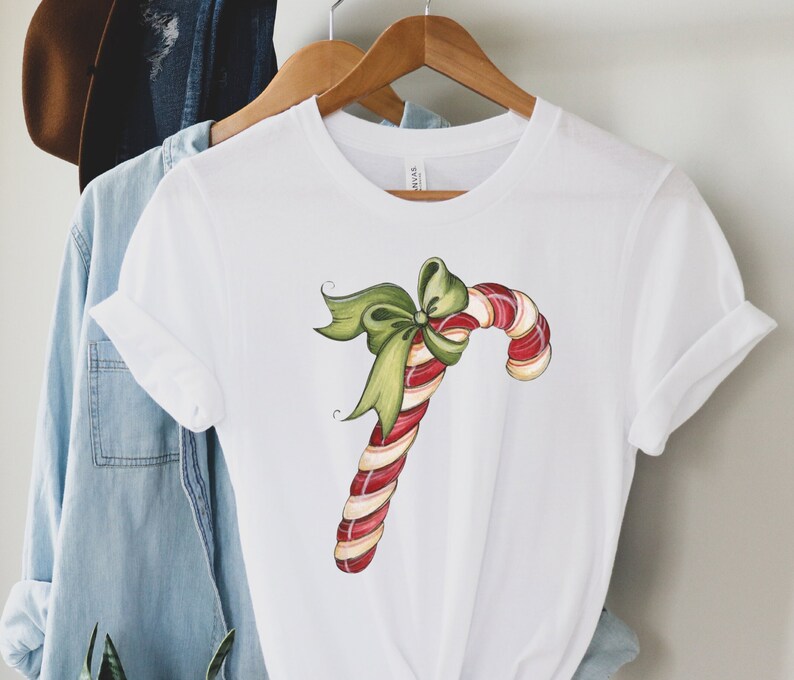 May include: White t-shirt with a red and white candy cane with a green bow on top. The candy cane is drawn in a cartoon style.