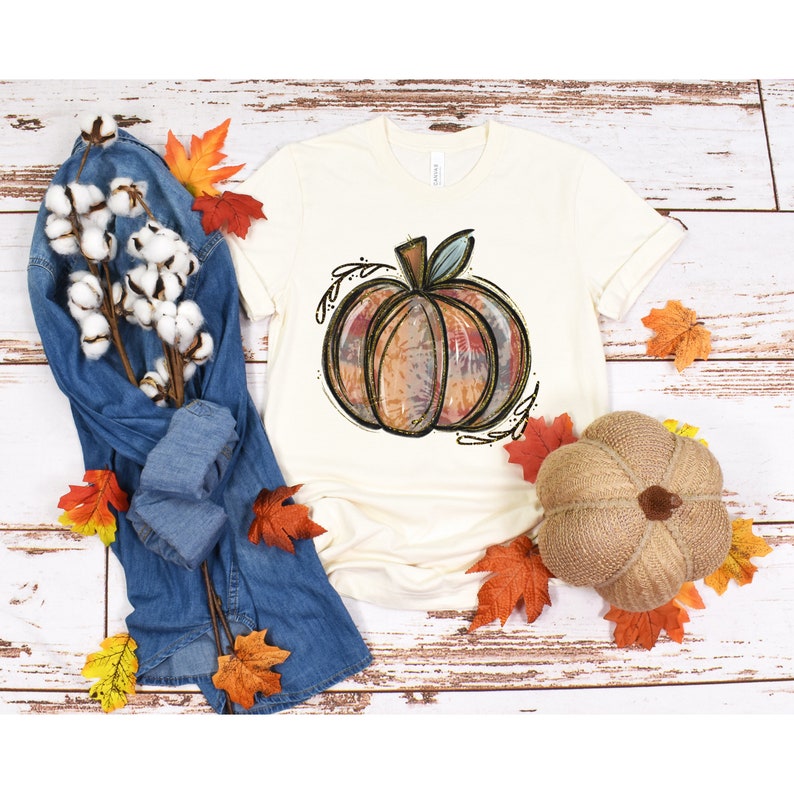 May include: A white t-shirt with a hand-drawn illustration of a pumpkin in shades of orange, brown, and blue. The pumpkin is surrounded by leaves and vines.