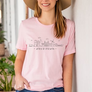 Może przedstawiać: Różowy t-shirt z czarnym rysunkiem linii horyzontu Amsterdamu, w tym wiatrak i tekst "AMSTERDAM".