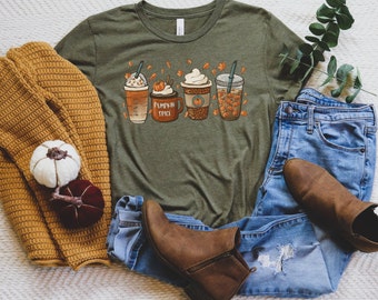 Puede incluir: Una camiseta verde con un gráfico de tres tazas de café con temática otoñal. Las tazas están decoradas con calabazas, hojas y el texto "Pumpkin Spice". La camiseta está colocada sobre una superficie blanca con un par de botines marrones, unos vaqueros azules y un suéter de punto marrón.