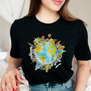 May include: A black t-shirt with a watercolor illustration of the Earth surrounded by iconic landmarks from around the world. The landmarks are drawn in a colorful style and include the Eiffel Tower, the Statue of Liberty, the Great Wall of China, and the Taj Mahal.