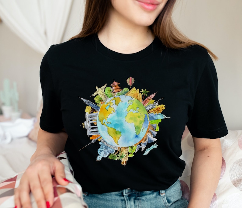 May include: A black t-shirt with a watercolor illustration of the Earth surrounded by iconic landmarks from around the world. The landmarks are drawn in a colorful style and include the Eiffel Tower, the Statue of Liberty, the Great Wall of China, and the Taj Mahal.