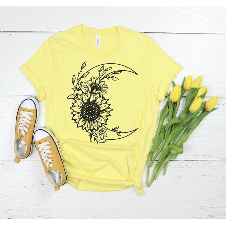 May include: A yellow t-shirt with a black graphic of a crescent moon with sunflowers and leaves.