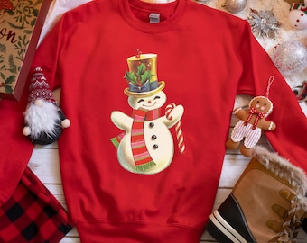 Op de afbeelding: Rood sweatshirt met een sneeuwpop-afbeelding die een hoge hoed, een sjaal draagt en een snoepstok vasthoudt. De sneeuwpop glimlacht en heeft een vrolijke uitdrukking.