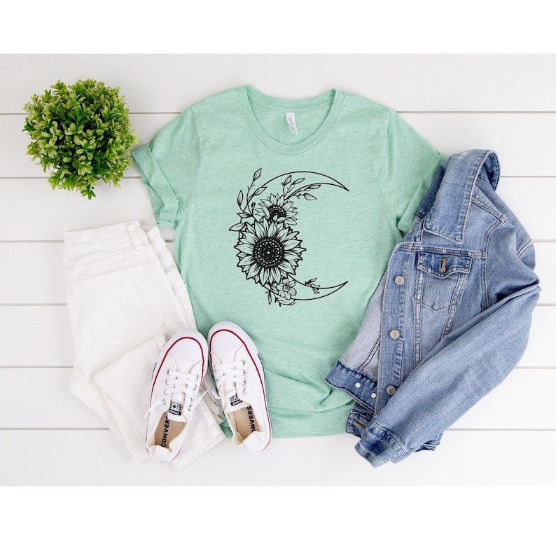 May include: A light green t-shirt with a black and white graphic of a crescent moon with sunflowers and leaves around it.