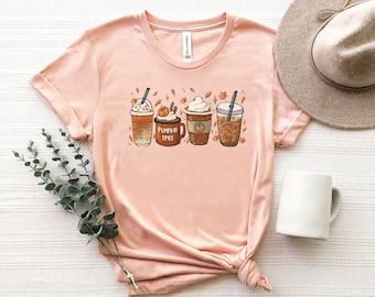 Puede incluir: Una camiseta de color melocotón con un gráfico de cuatro bebidas de café de temática otoñal. El texto "Pumpkin Spice" está impreso en la taza.