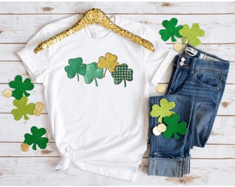 May include: White t-shirt with a green, gold, and black shamrock design. The shamrocks are glittery and have a checkered pattern.