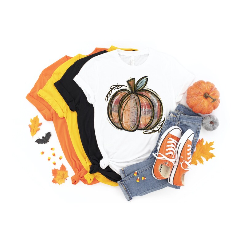 May include: A white t-shirt with a colorful, tie-dye style pumpkin graphic. The shirt is layered with other t-shirts in orange, yellow, and black. A pair of orange sneakers and blue jeans are also pictured.