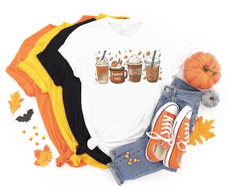 Puede incluir: Una camiseta blanca con un gráfico de cuatro bebidas de café con temática otoñal. Las bebidas están decoradas con hojas de otoño, palitos de canela y crema batida. El texto "Pumpkin Spice" está escrito en una de las tazas. La camiseta se muestra con un par de zapatillas naranjas, jeans y decoraciones de otoño.