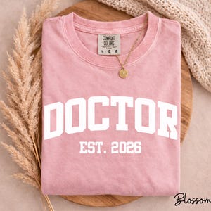 May include: A dusty rose-colored t-shirt with the word "DOCTOR" printed in large, white, block letters. Below the word "DOCTOR" is the text "EST. 2026" in white. A gold necklace with a circular pendant rests on the shirt.