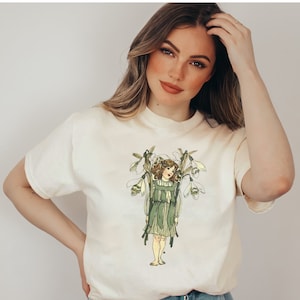 Puede incluir: Una camiseta blanca con una ilustración vintage de una niña con un vestido verde que sostiene un ramo de flores de campanilla de invierno blancas.