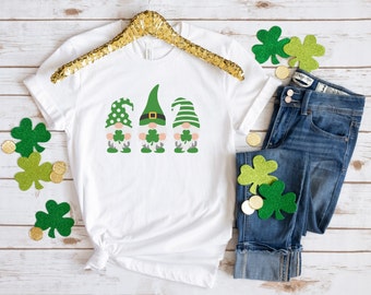May include: White t-shirt with three green gnomes wearing hats and holding shamrocks. The gnomes are in a row on the shirt. The shirt is on a white wooden background.