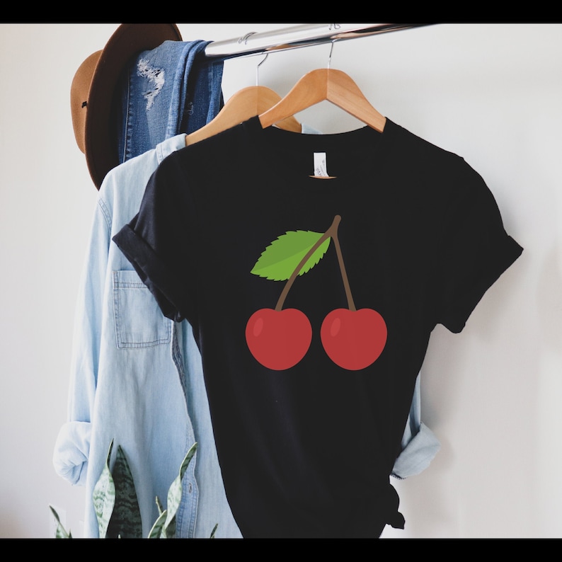 May include: Black t-shirt with a graphic of two red cherries and a green leaf. The t-shirt is hanging on a wooden hanger. Other clothing items are visible in the background. The t-shirt is made of a soft material.