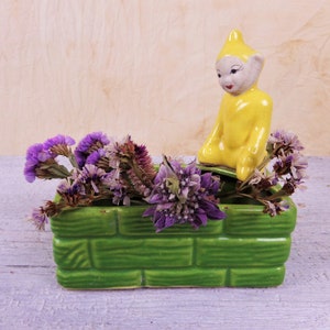 May include: A green ceramic planter with a brick pattern, filled with purple flowers and a yellow ceramic gnome figurine sitting on top.