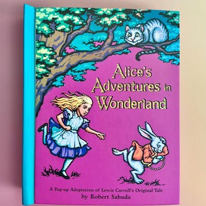 May include: A hardcover book titled "Alice's Adventures in Wonderland" with a pop-up adaptation by Robert Sabuda. The cover features illustrations of Alice, the White Rabbit, and the Cheshire Cat on a purple background.