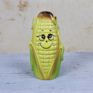 May include: A ceramic corn on the cob piggy bank with a smiling face and large eyes. The corn is yellow with green husks and brown silk.