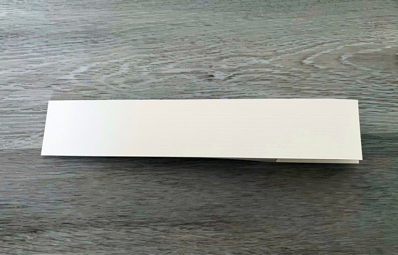 Puede incluir: Una pila de tarjetas blancas plegadas con una superficie texturizada. Las tarjetas son rectangulares y parecen estar en blanco, descansando sobre una superficie de madera gris. Las tarjetas son probablemente para invitaciones o notas.