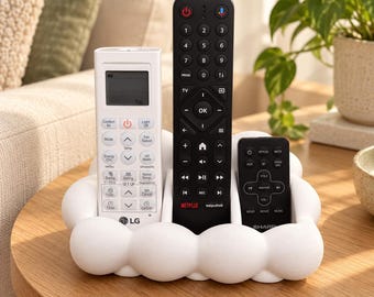 Puede incluir: Un soporte para mandos a distancia blanco con forma de nube y textura redondeada. Contiene tres mandos: un mando de aire acondicionado LG blanco a la izquierda, un mando de televisión negro alto en el centro y un mando Sharp negro pequeño.