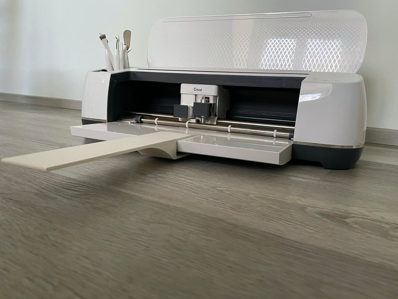 Puede incluir: M&aacute;quina de corte Cricut blanca y gris con la tapa abierta, que revela la cuchilla de corte y los componentes internos. La m&aacute;quina tiene una bandeja extra&iacute;ble y un compartimento para herramientas. El logotipo de Cricut es visible.