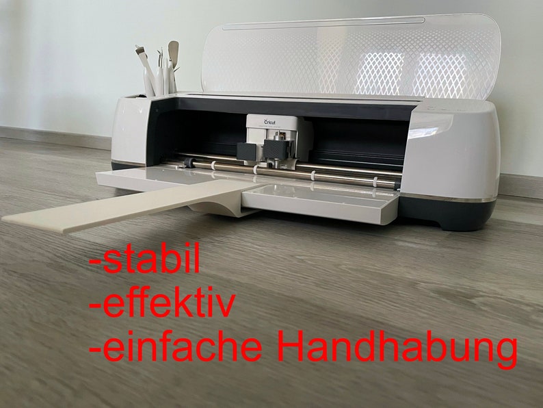 Puede incluir: M&aacute;quina de corte Cricut blanca y gris con la tapa abierta, mostrando la cuchilla y el &aacute;rea de corte. La m&aacute;quina tiene una bandeja extendida y un compartimento para herramientas. El texto rojo dice: "-estable -efectivo -f&aacute;cil de usar."