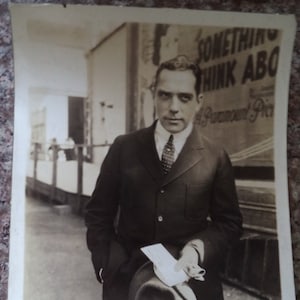 May include: A black and white photograph of a man wearing a suit and tie. He is holding a letter in his right hand and a hat in his left hand. The photo was taken in front of a building with a sign that reads "Something Think Abo Paramount Pic."