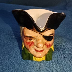 Op de afbeelding: Een keramische creamer in de vorm van een piratenhoofd. De creamer heeft een witte hoed met zwarte rand, een zwart ooglapje en een geschilderd gezicht met rode wangen en een gele en groene kraag.