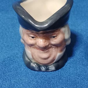 Può includere: Una piccola crema in ceramica a forma di testa d'uomo. La crema ha un interno bianco ed è dipinta con un cappello grigio, una parrucca bianca e un viso sorridente con guance rosee. Sfondo blu.