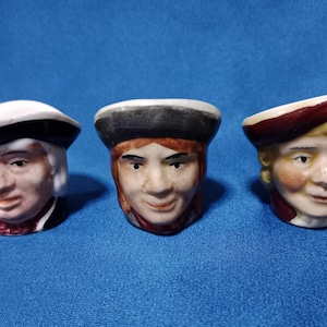 Puede incluir: Tres figuras de cerámica con forma de cabezas humanas, cada una con un sombrero. La primera tiene pelo blanco y un sombrero blanco con ala negra. La segunda tiene pelo castaño y un sombrero negro. La tercera tiene pelo rubio y un sombrero granate.