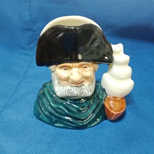 Puede incluir: Una taza de cerámica con forma de anciano con barba blanca y sombrero negro. La taza tiene un chal verde y un diseño blanco apilado en el lateral. La taza está sobre una superficie azul.