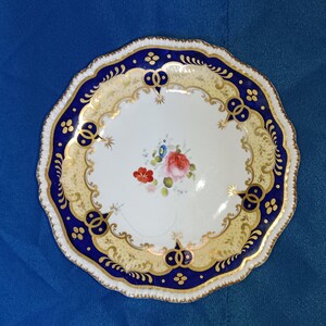Pode incluir: Um prato de porcelana com borda ondulada. O centro branco apresenta um desenho floral com flores vermelhas, rosas e azuis. A borda azul e dourada apresenta detalhes dourados intrincados. Um objeto decorativo.